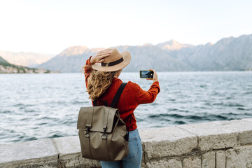 Back view of young woman with backpack using smartphone to take photo of mountains and lake while standing near pier railing. Lifestyle, travel, tourism, active life.