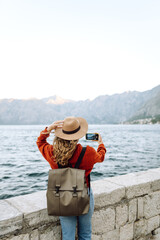 Back view of young woman with backpack using smartphone to take photo of mountains and lake while...