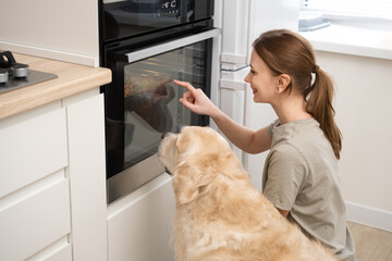 A young woman looks into the oven with her dog. A young woman waiting for pizza to be cooked with her pet
