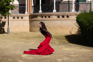 Beautiful woman dancing flamenco in Seville, Spain. She is wearing a red and black gypsy dress and dancing flamenco with a lot of art. In the background a monument with arches and columns. - obrazy, fototapety, plakaty