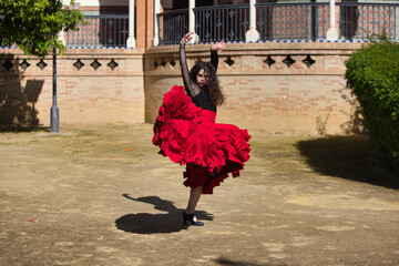 Beautiful woman dancing flamenco in Seville, Spain. She wears a red and black dress typical of a flamenco dancer with a lot of art, you can see the movement in the air of the frilly dress. - obrazy, fototapety, plakaty