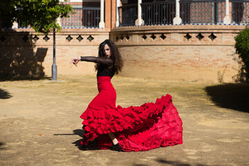 Beautiful woman dancing flamenco in Seville, Spain. She wears a red and black dress typical of a flamenco dancer with a lot of art, you can see the movement in the air of the frilly dress. - obrazy, fototapety, plakaty