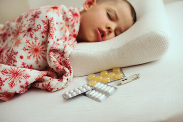 Sick little boy sleeping in white bed with a thermometer and antipyretic tablets on a orthopedic pillow
- 790318224
