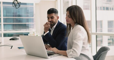 Indian and Latina colleagues, business partners met in boardroom looking at laptop screen, reviewing trends, discuss marketing campaign revenue, feel satisfied with sales results, engaged in teamwork