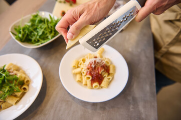 Directly above chef hands using grater, grating cheese on freshly cooked pasta with tomato sauce, seasoning and plating up the meal before serving it