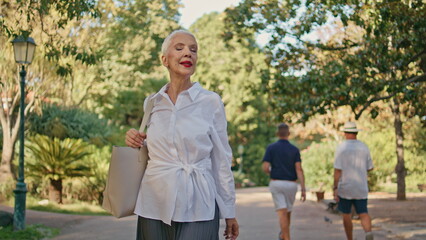 Elderly woman going park path in sunlight. Smiling relaxed senior resting alone
