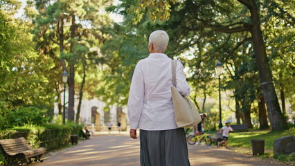 Older woman enjoying stroll in green park back view. Relaxed senior rest alone