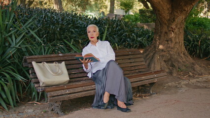 Pensioner relaxing city park holding book. Smiling elegant woman enjoying day