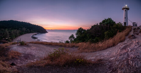The sunset casts a warm glow over a lighthouse and a quiet beach nestled between lush hills,...