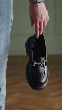 Hand pointing at a black loafer on wooden floor with a partial view of a person wearing jeans. Conceptual shot for shoe features with focus on design and style.