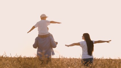 Happy family walking on dry wheat field flying with open hands playing together back view. Cheerful mother father and son fooling relaxing enjoy freedom at sunny sunset harvest natural landscape - obrazy, fototapety, plakaty