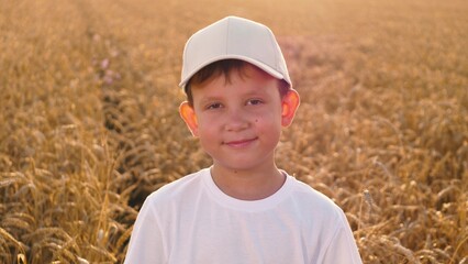 Naklejka na ściany i meble Portrait adorable little boy child smiling posing at autumn dry wheat field closeup. Happy male kid in white cap and shirt relaxing outdoor natural sunny sunlight surrounded by cereal harvest meadow