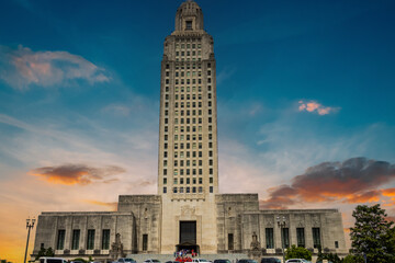 The Louisiana State Capitol building with lush green trees and plants, stone statues, people standing on the stairs and powerful clouds at sunset in Baton Rouge Louisiana USA