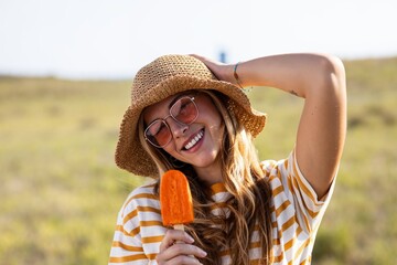 Attractive young woman eating a orange popsicle looking at the camera on summer - 790400410