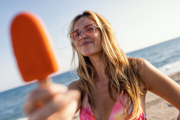 Attractive young woman in bikini eating a orange popsicle looking at the camera on summer - 790400692