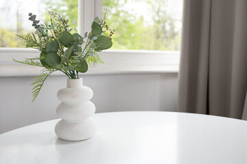 Background of white vase with artificial greenery on white table and window at the back