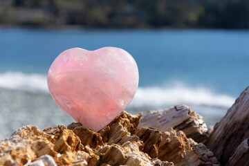 A close up image of a rose quartz crystal heart resting on driftwood with the Pacific Ocean waves in the background. 