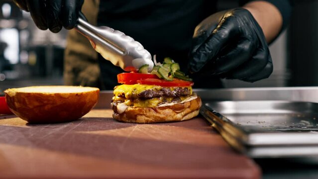 A chef in a professional kitchen places cucumber slices on beef cutlet with tongs