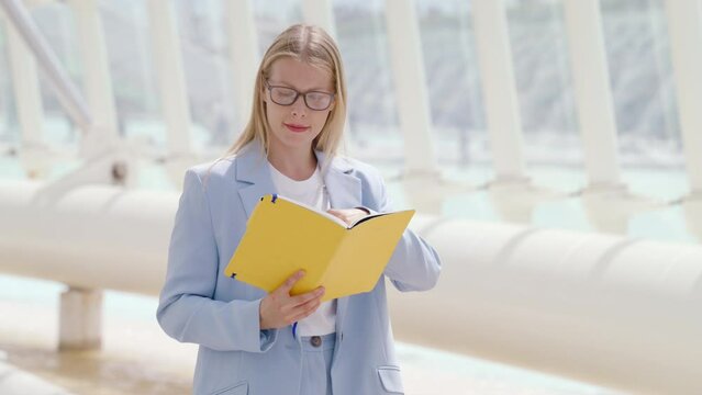 Focused young businesswoman in glasses and a light blue suit reading a yellow document, with white architectural columns in the background. The scene depicts serious work outdoors. Slow motion. 