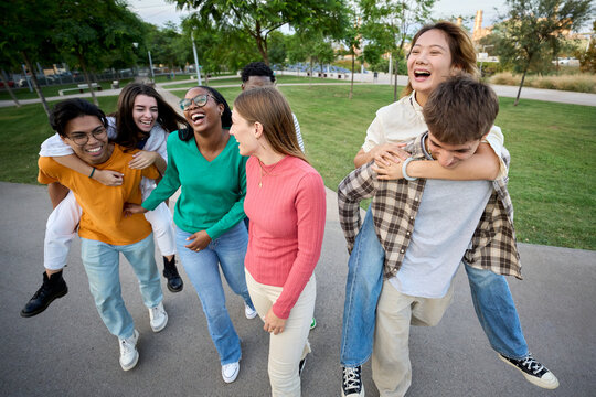 A large multiracial group of teenagers laughing and having fun piggybacking together in an outdoor garden area. Young students enjoying their leisure and free time with friends in the park