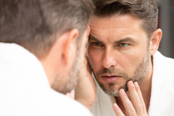 a man in a white bathrobe in the bathroom looking at the skin on his faceSpa male model, skincare product for skin face. Skin care. Male face cream. Man with perfect soft skin. Beauty cosmetic product