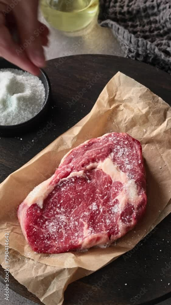 Wall mural chef sprinkles salt on fresh raw beef steak in kitchen, close-up