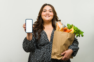 Cheerful plus-size woman holding a grocery bag and displaying a blank smartphone screen in a studio setting - 790415841