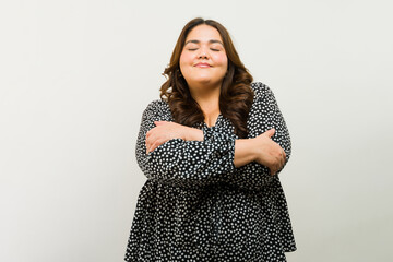 Plus-size woman with a content smile hugging her self and showing self-love in a studio - 790416004