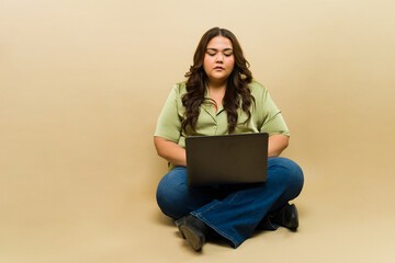 Thoughtful plus-size woman using a laptop on a beige background in a studio setting