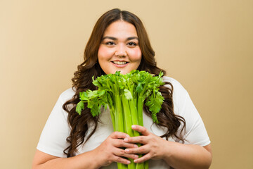 Happy curvy woman with a bright smile posing in a studio with a bunch of fresh celery sticks - 790416447