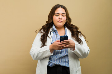 Plus-size female doctor focuses on her phone, exuding confidence while wearing lab coat in a studio