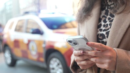 Unrecognizable young woman requesting urgent assistance in the street next to emergency car. Close-up of a woman's hands holding and using her mobile phone.