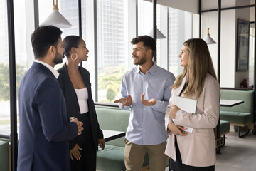 Positive young Arab professional man talking to diverse colleagues in office hall, speaking, smiling, standing in group of multiethnic managers in formal suits, discussing teamwork, communication