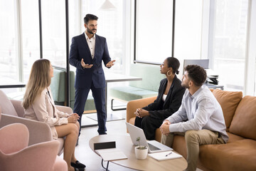 Ambitious confident young boss talking to diverse team of project managers, explaining work plan, marketing strategy, instructing employees, standing in office co-working space