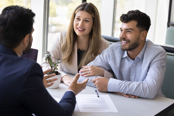 Happy young diverse husband and wife consulting legal adviser, realtor, talking, listening to consultant at meeting, sitting at table with paper document, discussing loan mortgage agreement