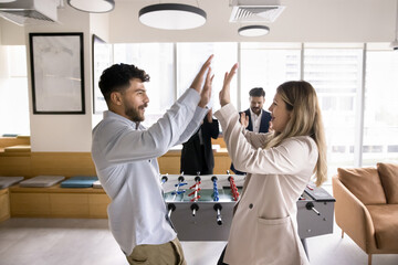 Happy couple of office workers beating colleagues in toy football, giving high five at table soccer stand, smiling, laughing, enjoying win, team success, recreation on work break