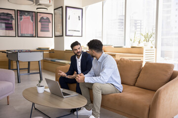 Young business startup leader man consulting Indian professional expert in formal suit. Colleagues meeting on sofa, talking on work break in office hall, sitting at laptop