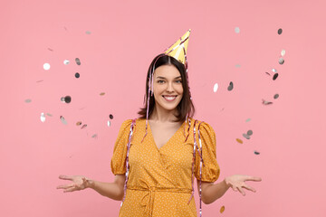 Happy young woman in party hat near flying confetti on pink background
