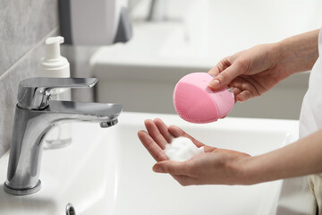 Washing face. Woman with brush and cleansing foam above sink in bathroom, closeup