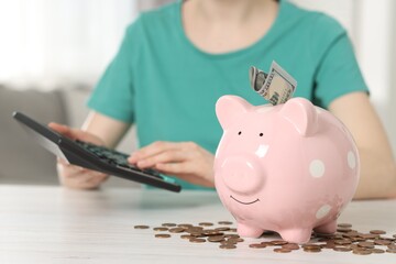 Financial savings. Woman using calculator at white wooden table indoors, focus on piggy bank and...