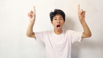 young asian man poses surprised while pointing upwards on a white background