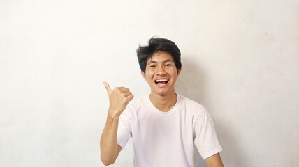Young Asian man posing happily while pointing to the side using his thumb on a white background