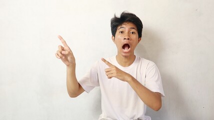young asian man poses surprised while pointing upwards on a white background