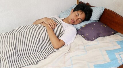 A young Asian man in a white shirt is sleeping on his bedroom mattress