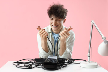 Electrocuted young man with burnt face and jumper cables at table on pink background