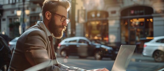 Bearded man sitting on a bench in a bustling city street while focused on his laptop screen - obrazy, fototapety, plakaty