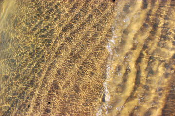 Close-up of a river bed with yellow sand, stones curling from the wave, in bright sunlight with beautiful highlights on the water. Texture of river sand. Texture with seashells and pebbles on the wet 