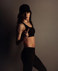 Fuck you. Sport sexy muscular woman posing in black sport bra, cap and showing the fuck sign the hand, standing on dark shadow studio background. Front body view. Sporty lifestyle. - 790498261