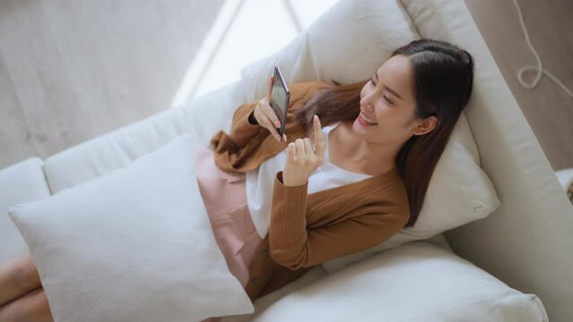 High angle view of happy cheerful positive young Asian woman on the phone at home, smiling girl using app and browsing on the internet on smartphone lying on sofa