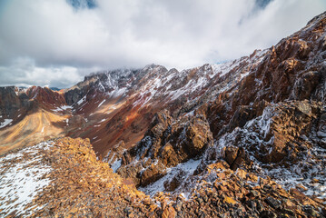 Scenic top view from precipice edge to multicolor big mountain in freshly fallen snow in sunlight in low clouds. Large multi-color snowy rocky ridge in cloudy sky. Snow-covered sharp rocks above abyss - 790538221